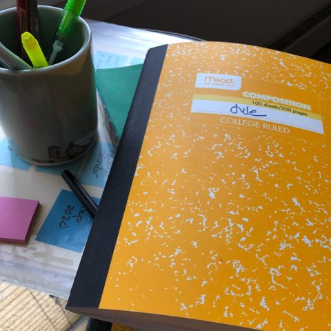 A yellow composition notebook on a table, with a cup of pens next to it