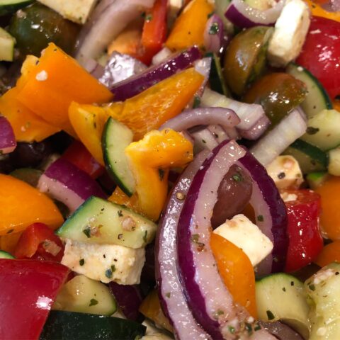Close up of a Greek salad, shiny with olive oil and colorful with red and orange peppers, white feta cheese, green cucumbers, dark green olives, and purple onions
