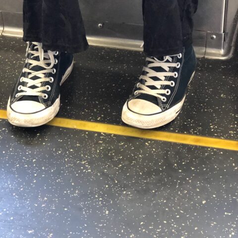 Black converse sneakers with white laces and toes on a train, the floor black and speckled with white.
