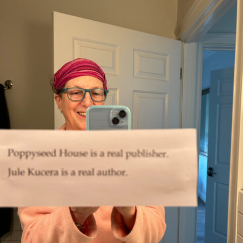 Jule Kucera taking a selfie in a mirror, with a note taped to the mirror: "Poppyseed House is a real publisher. Jule Kucera is a real author."