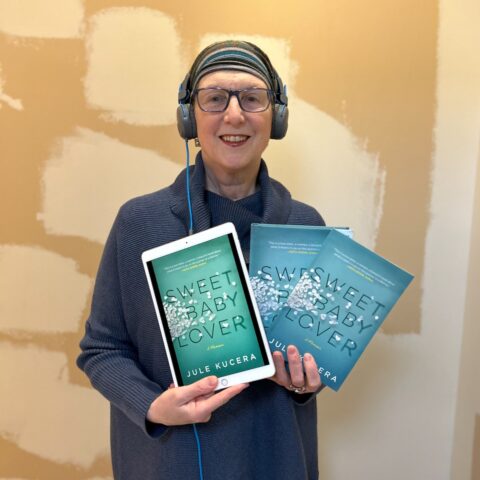 Jule Kucera holding an iPad showing Sweet Baby Lover, plus the large print book and paperback book, and wearing headphones